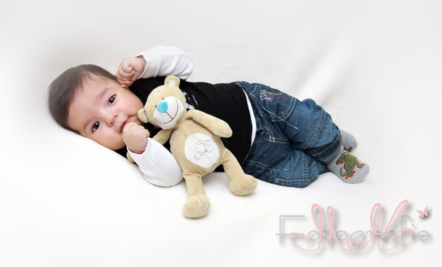 Die Fotografie eines Babys, es liegt auf der Seite und lächelt mit offenen Augen in die Kamera, ein hellbrauner Teddybär liegt vor ihm. Das Baby hat eine blaue Jeanshose an und ein schwarzes T-shirt mit Aufdruck und weißen Ärmeln. 