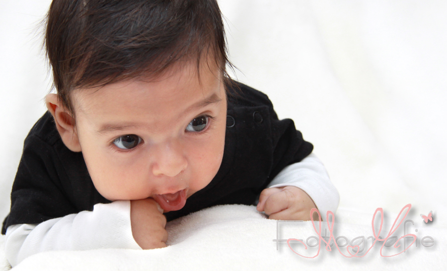Babyfotografie. Das Baby liegt auf dem Bauch, lächelt und schaut mit großen Augen in die Welt.
