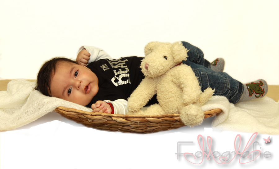 Auf einer weißen gestrickten Decke liegt das zwei Monate alte Baby in einem runden hellbraunen Korb. Farbfoto.