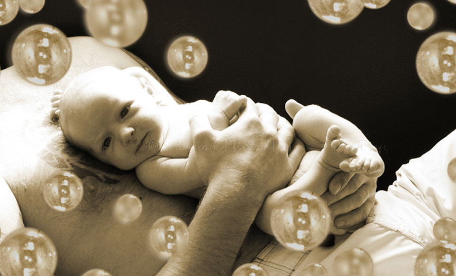 Babyfoto in Sepia. Ein neugeborenes Kind liegt auf dem Bauch seines Vaters, darum herum sind Seifenblasen